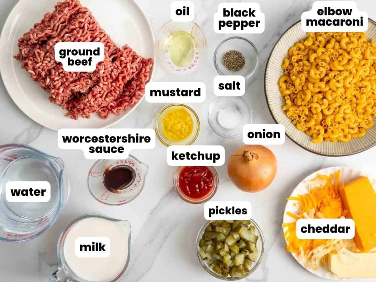 Ingredients for cheeseburger mac and cheese, including elbow macaroni, ground beef, and pickles, arranged on a marble countertop.