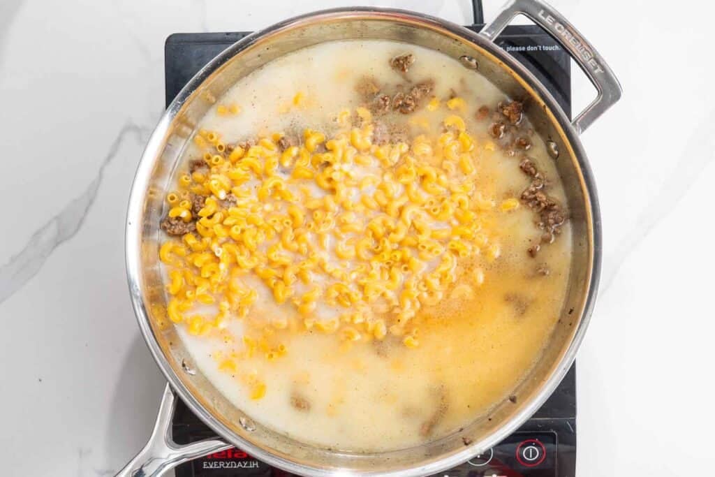 Water, milk, and macaroni added to a skillet of cooked ground beef.