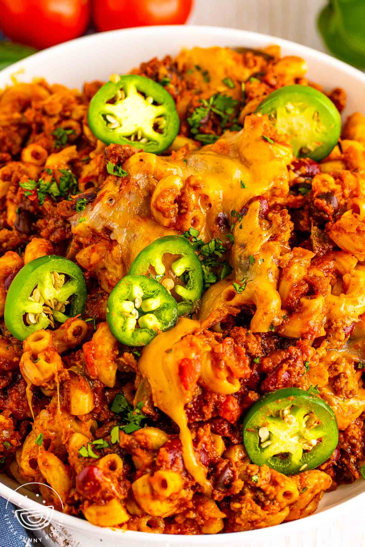 Bowl of chili macaroni with ground beef, beans, melted cheese, sliced jalapeños, and chopped cilantro.