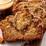 slices of banana bread on a wooden board next to a cup of coffee and in front of a bunch of bananas.