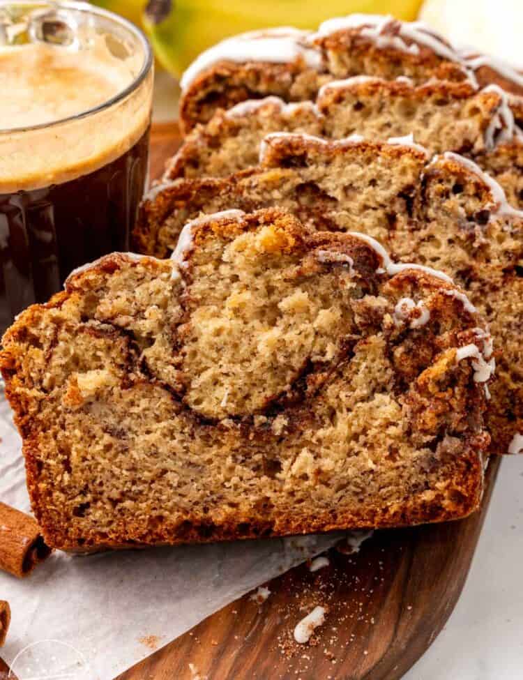 slices of banana bread on a wooden board next to a cup of coffee and in front of a bunch of bananas.