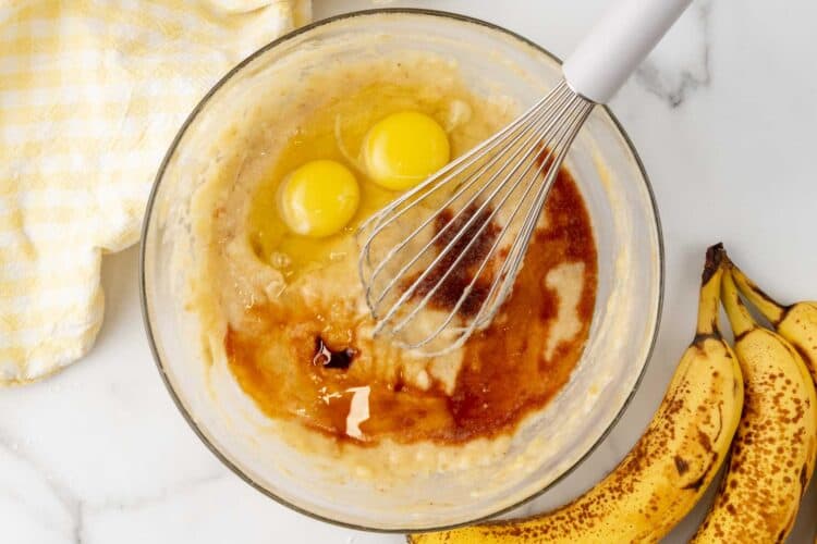 eggs, vanilla, and mashed bananas whisked together in a glass bowl. Next to the bowl are ripe bananas and a yellow and white gingham towel.