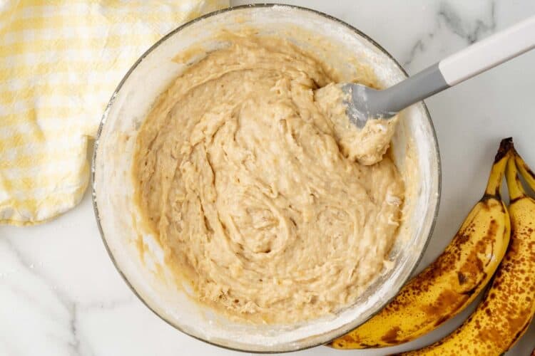 banana bread batter in a glass mixing bowl, stirred with a spatula. Next to the bowl is a bunch of ripe bananas.