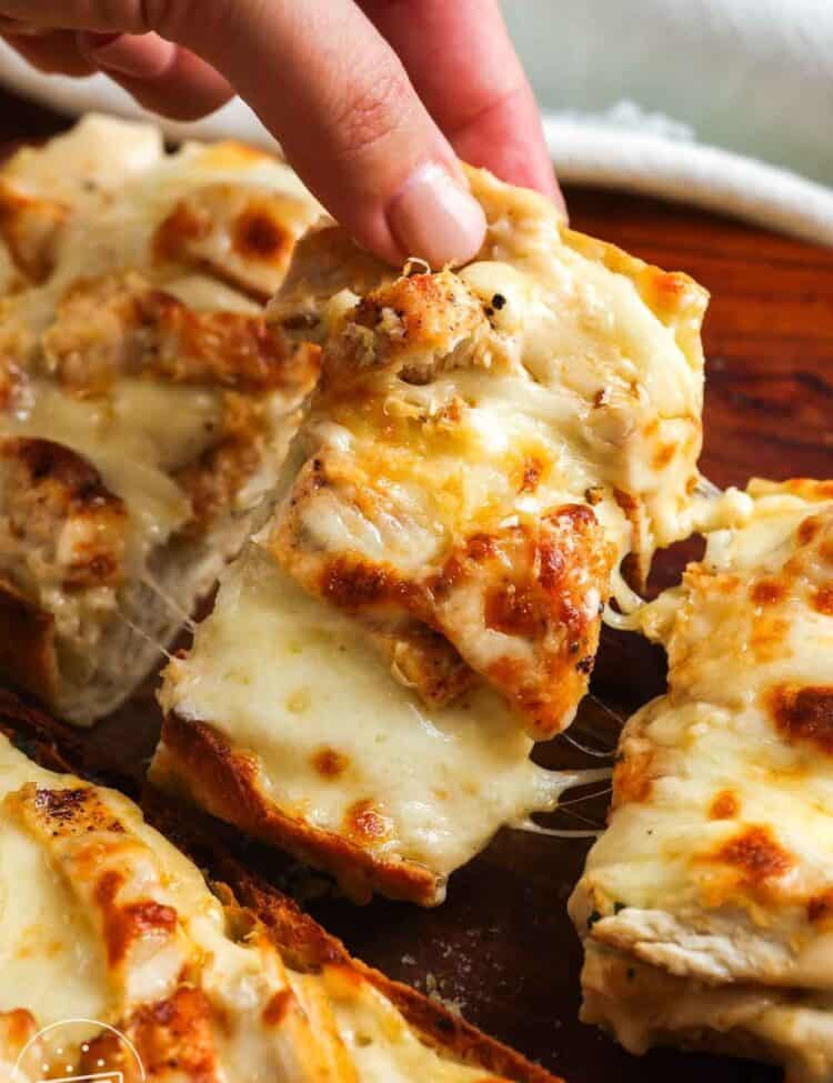 Hand picking up a cheesy slice of chicken Alfredo garlic bread with melted mozzarella and golden ciabatta.