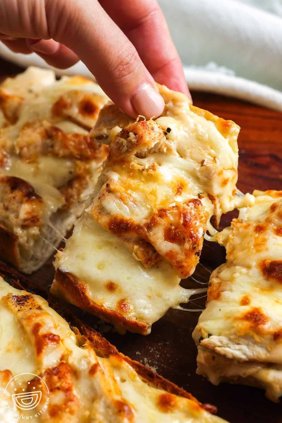 Hand picking up a cheesy slice of chicken Alfredo garlic bread with melted mozzarella and golden ciabatta.