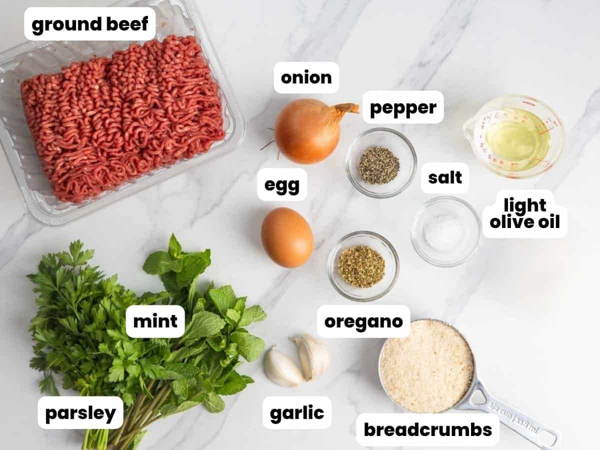 a pound of ground beef on a counter next to a bunch of fresh herbs, two garlic cloves, a cup of breadcrumbs, an egg, an onion, and seasonings.
