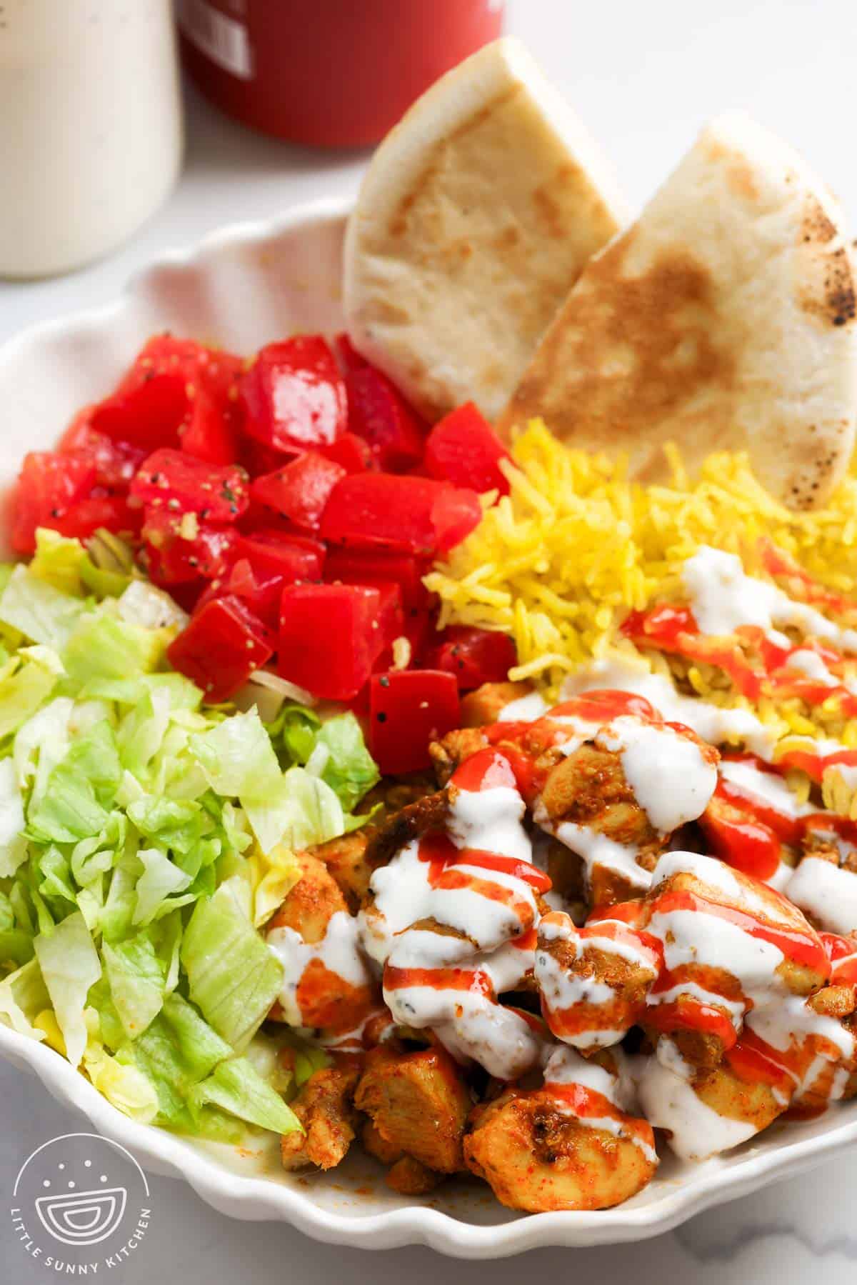 a plate of halal cart style chicken drizzled with white sauce and hot sauce, served with rice, pita, lettuce, and tomatoes.