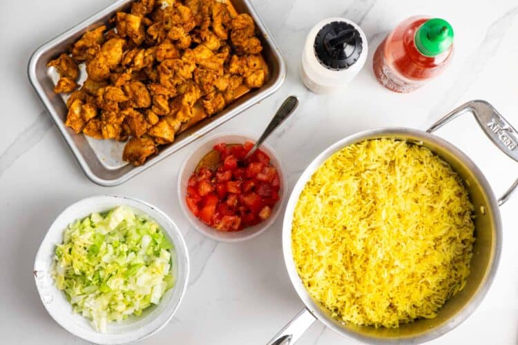 Components of Halal cart chicken rice bowls, before serving. There is a pot of yellow rice, a pan of cooked chicken, a bowl of seasoned tomatoes, a bowl of rice, a bottle of hot sauce, and a squeeze bottle of white sauce.