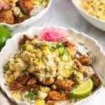 A bowl of rice topped with seasoned chicken and mexican street corn salad. On the side are lime wedges and pickled onions.