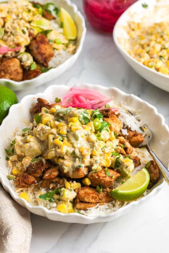 A bowl of rice topped with seasoned chicken and mexican street corn salad. On the side are lime wedges and pickled onions.