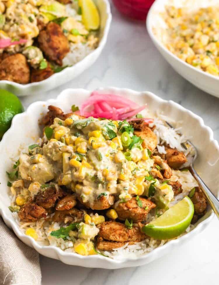 A bowl of rice topped with seasoned chicken and mexican street corn salad. On the side are lime wedges and pickled onions.