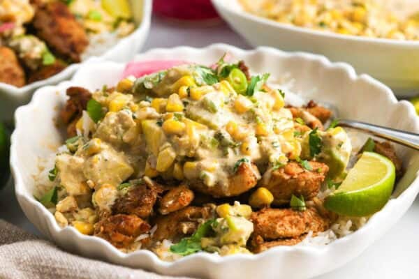 A white ceramic bowl of rice, Mexican seasoned chicken, and creamy street corn salad.