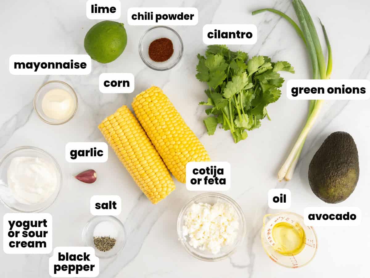Ingredients for street corn salad including fresh corn, cotija cheese, greek yogurt, limes, and avocado.