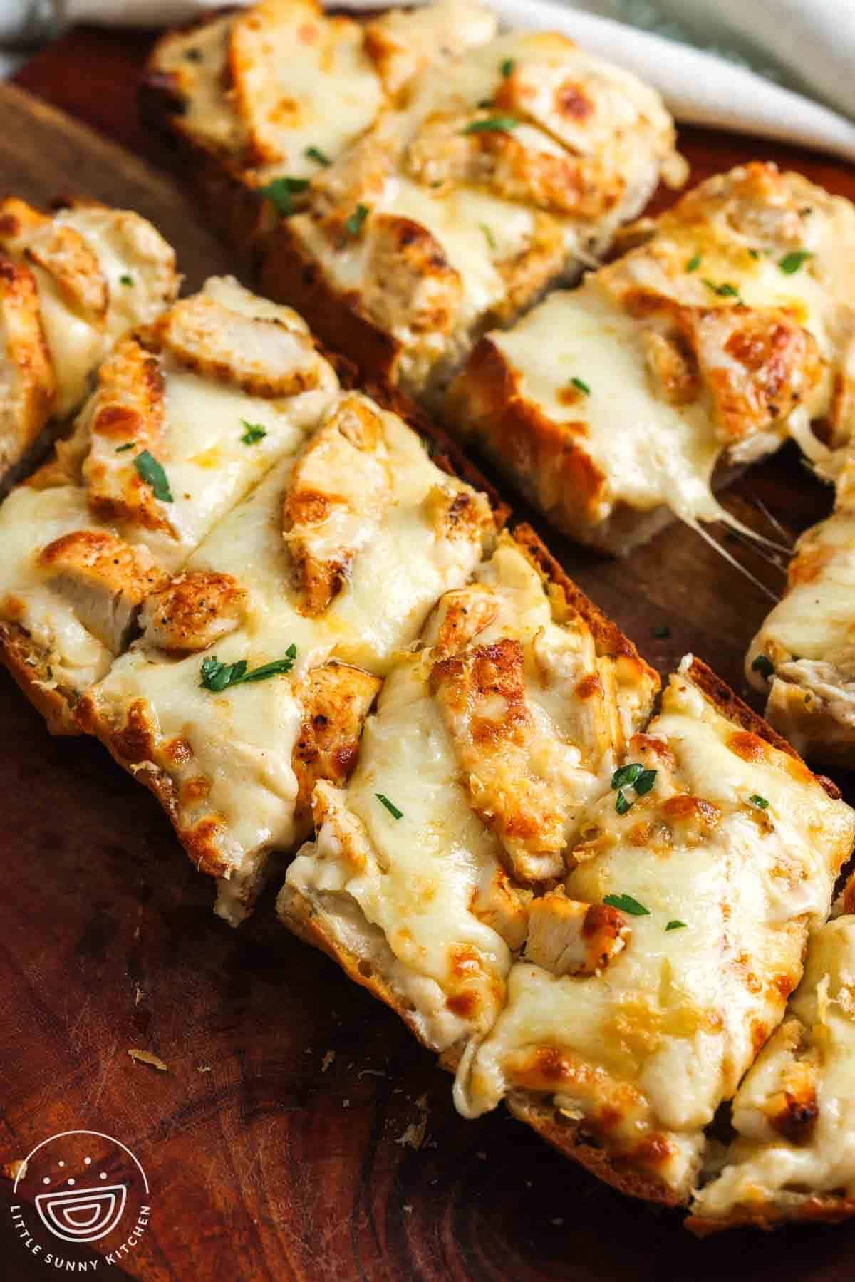 Sliced chicken Alfredo garlic bread with melted mozzarella, pieces of chicken, parsley, and a cheese pull on a wooden board.