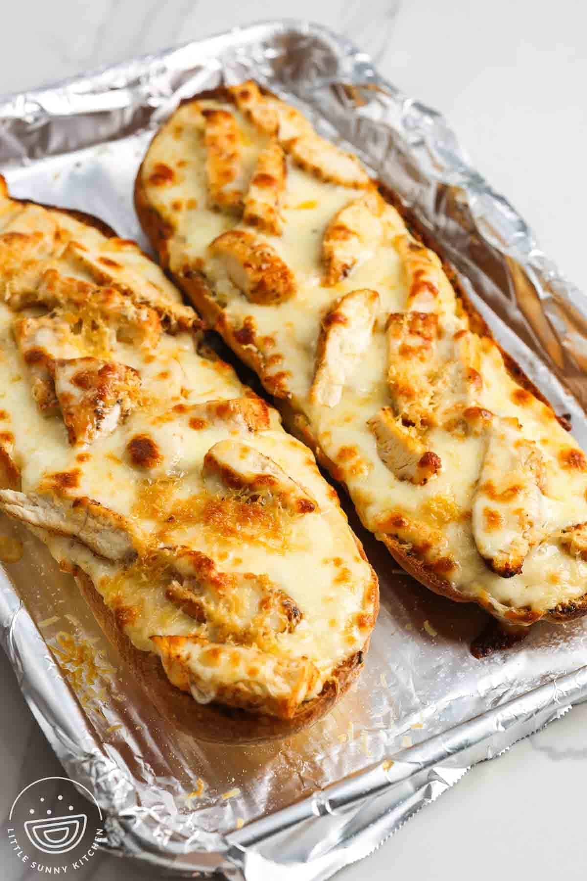 Two halves of chicken Alfredo garlic bread topped with melted mozzarella and sliced chicken on a foil-lined baking sheet.