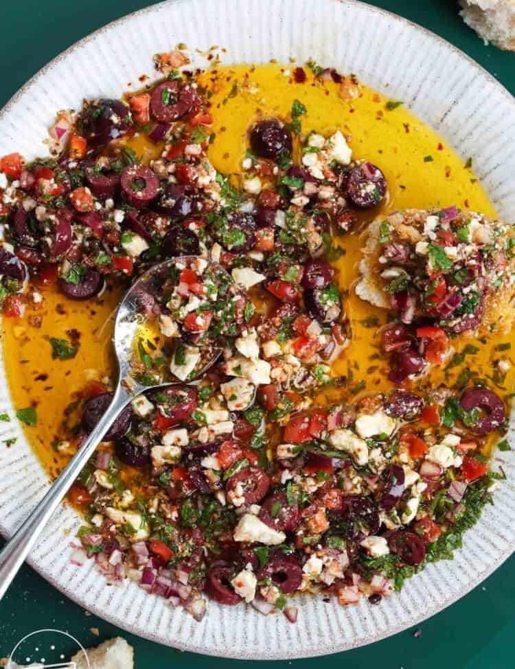 Mediterranean olive oil dip with olives, feta, tomatoes, herbs, and crusty bread in a shallow bowl.