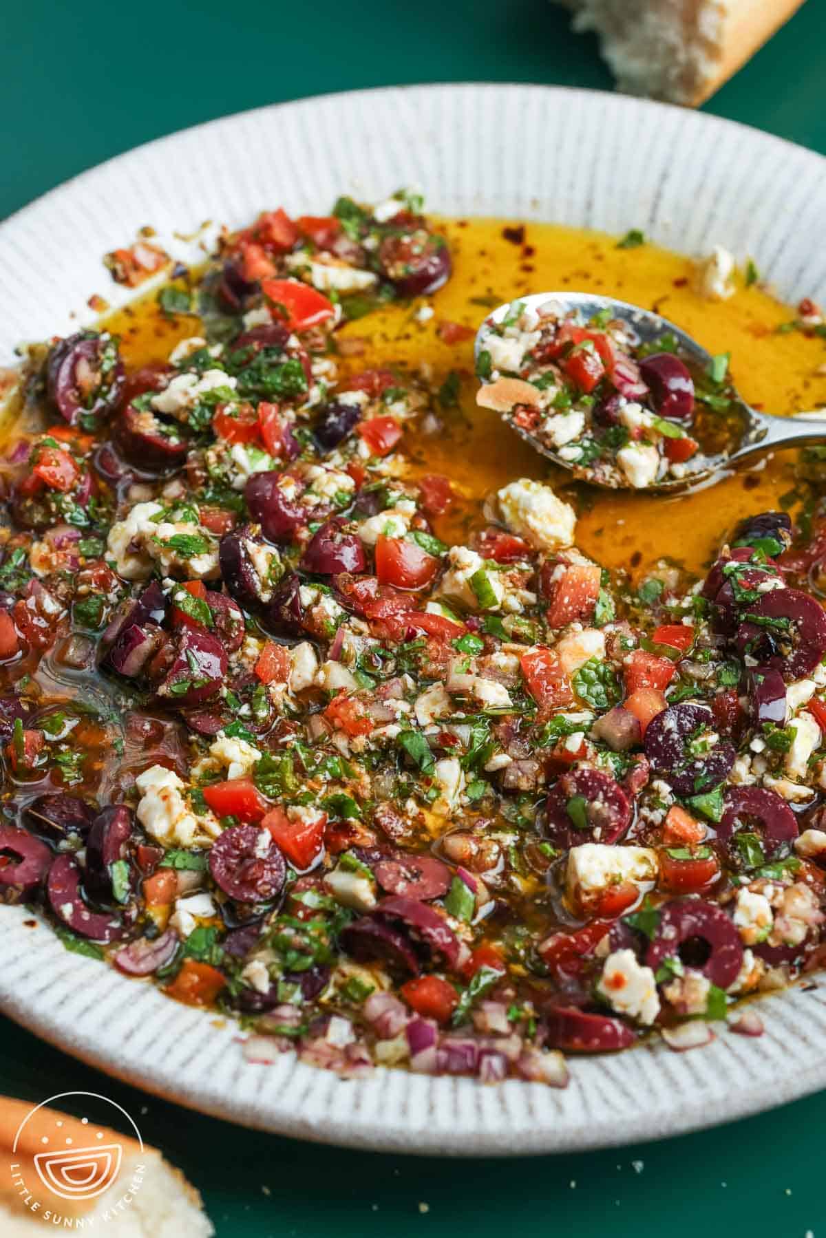 Spoonful of Mediterranean olive oil dip with olives, feta, tomatoes, and herbs in a shallow bowl.