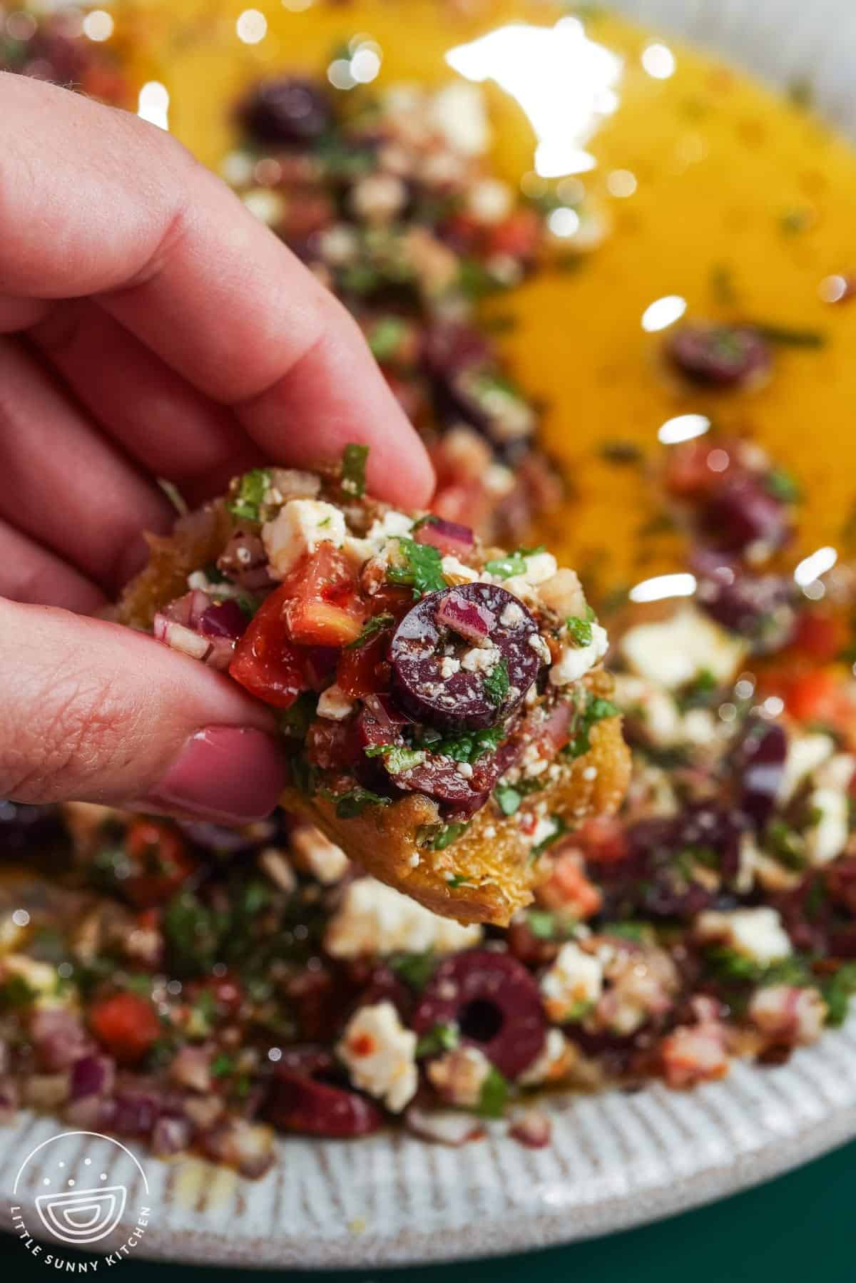 Hand holding crusty bread topped with Mediterranean olive oil dip, olives, feta, tomatoes, and herbs.
