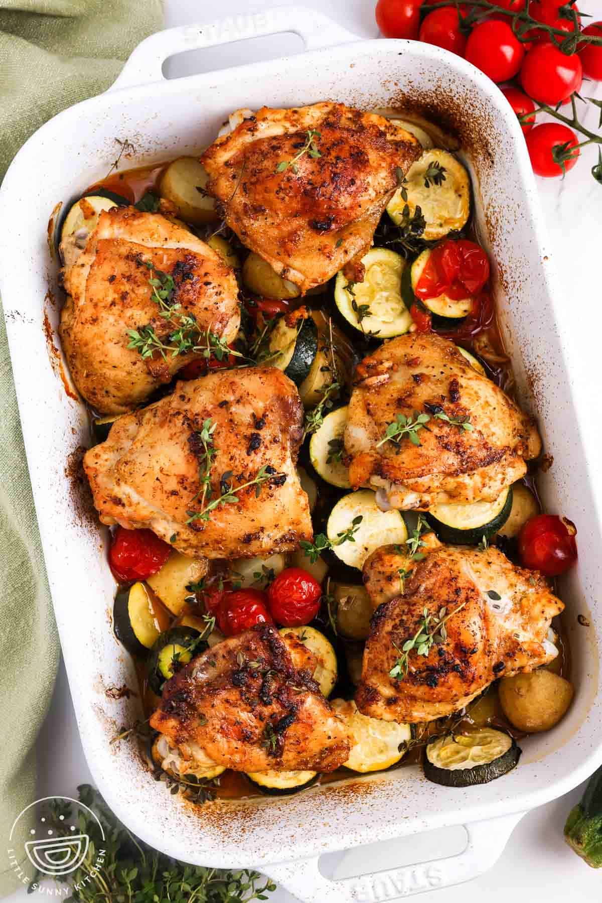 Overhead view of baked chicken thighs with zucchini, potatoes, tomatoes, and thyme in a white baking dish.