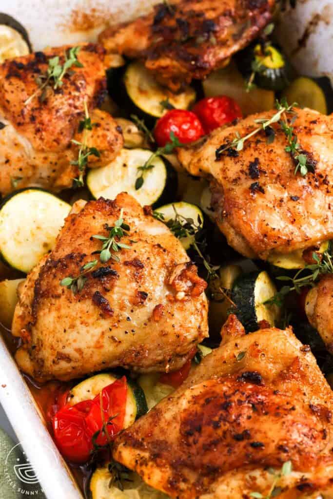 Close up of golden baked chicken thighs with zucchini, potatoes, tomatoes, and thyme.