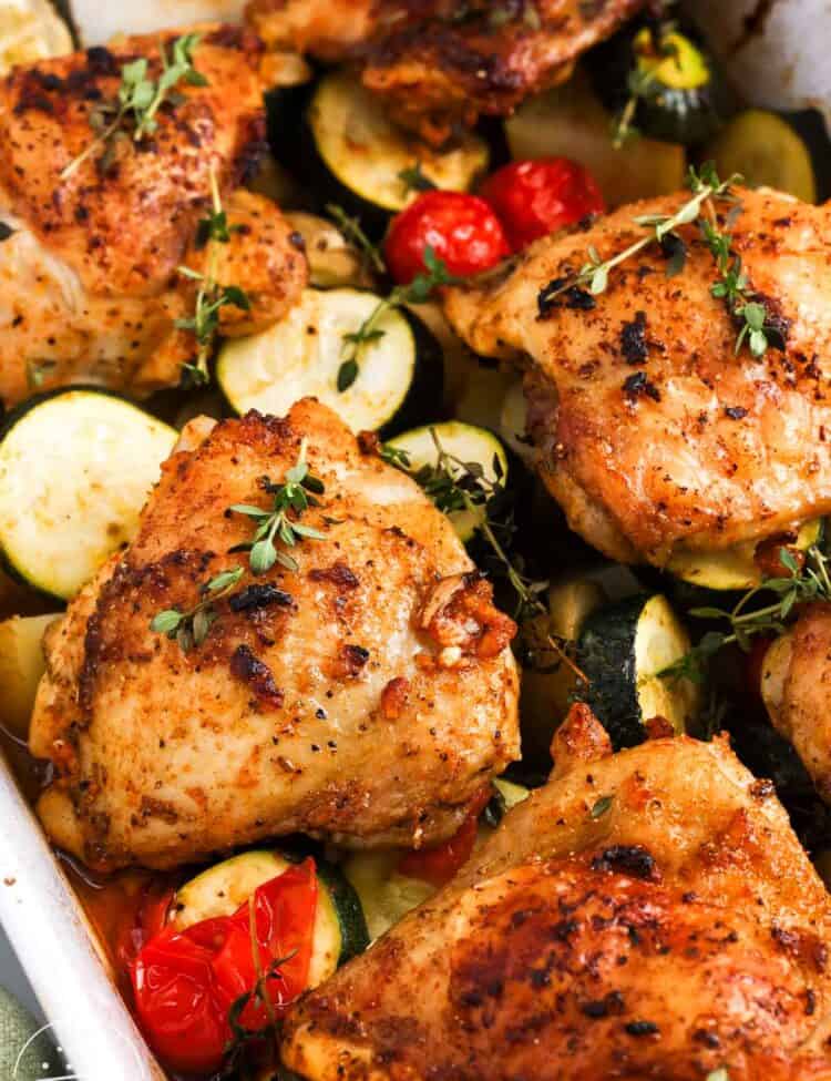 Close up of golden baked chicken thighs with zucchini, potatoes, tomatoes, and thyme.