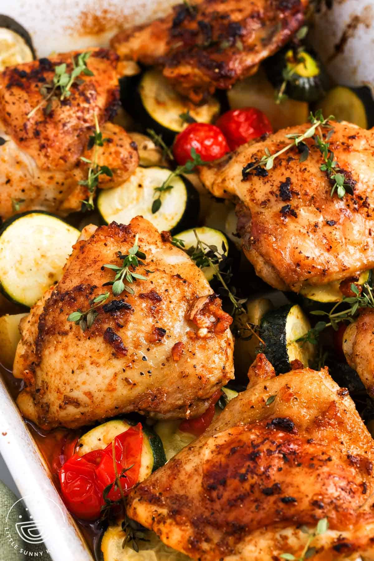 Baked chicken thighs with golden skin over zucchini, potatoes, and burst cherry tomatoes