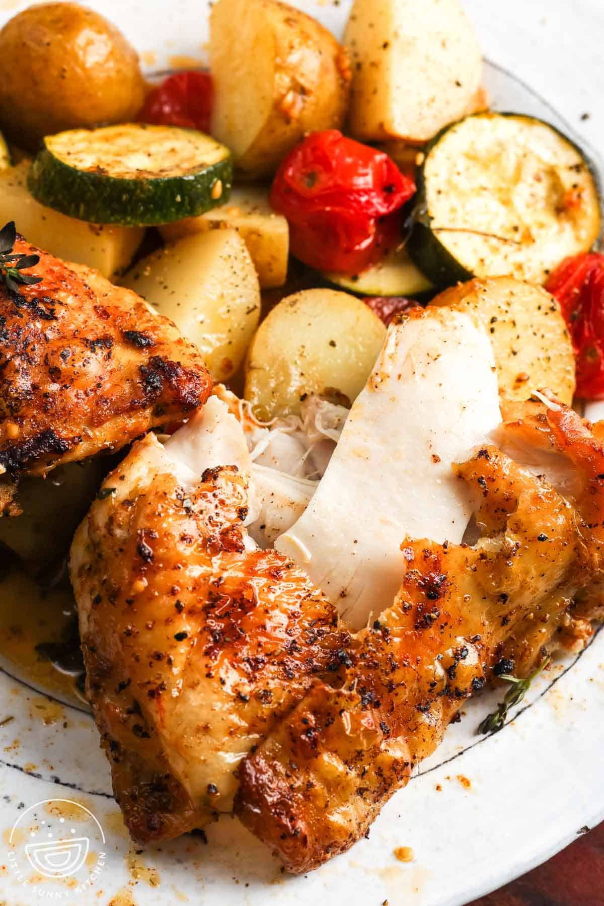 A serving of baked chicken thigh with roasted potatoes, zucchini, and tomatoes on a plate.