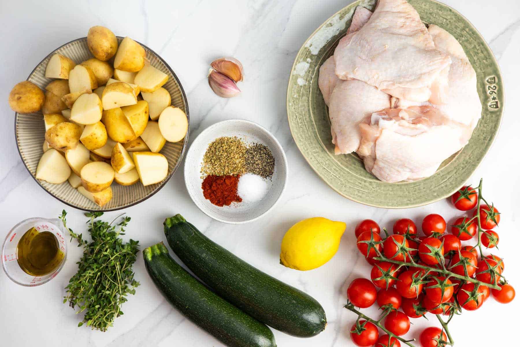 Ingredients for baked chicken thighs with summer vegetables, including chicken thighs, potatoes, zucchini, tomatoes, lemon, garlic, thyme, olive oil, and seasonings.