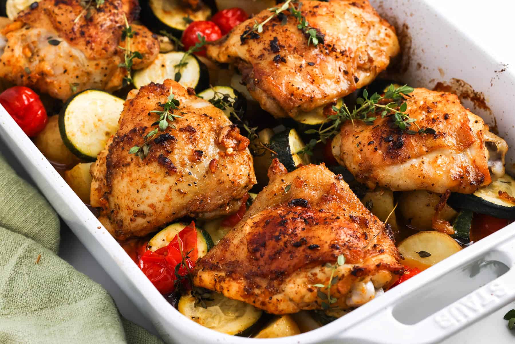 Baked chicken thighs with potatoes, zucchini, tomatoes, and thyme in a white baking dish.