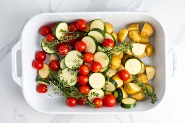 Potatoes, zucchini, cherry tomatoes, and thyme arranged in a white baking dish.
