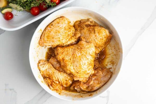 Chicken thighs coated in lemon garlic seasoning in a mixing bowl beside the prepared vegetables.