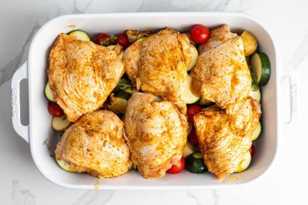 Seasoned chicken thighs arranged over potatoes, zucchini, tomatoes, and thyme before baking.