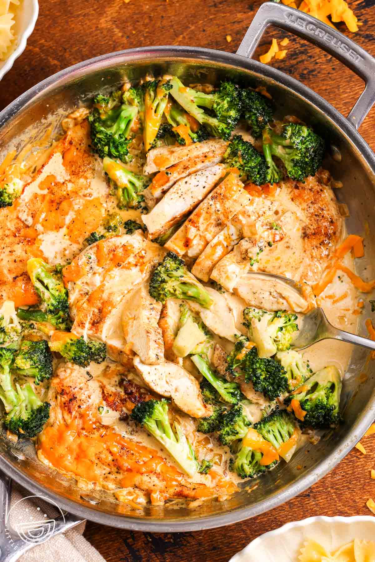 Creamy broccoli cheddar chicken in a skillet. next to the skillet are bowls of pasta.