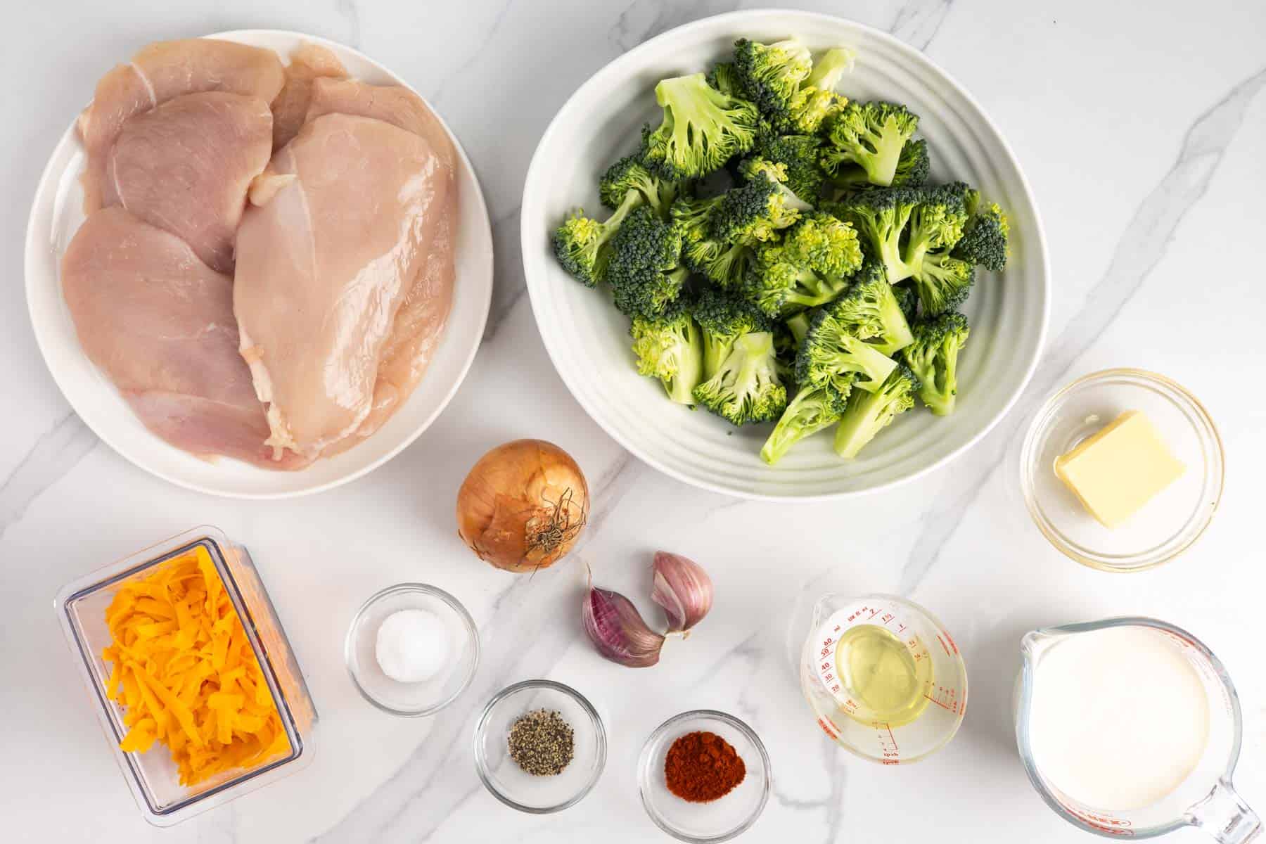 A plate of raw chicken cutlets next to a bowl of fresh broccoli florets and the ingredients to create a cheese sauce.