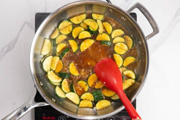 a sauce added to sauteed zucchini in a skillet, stirred with a red spoon.