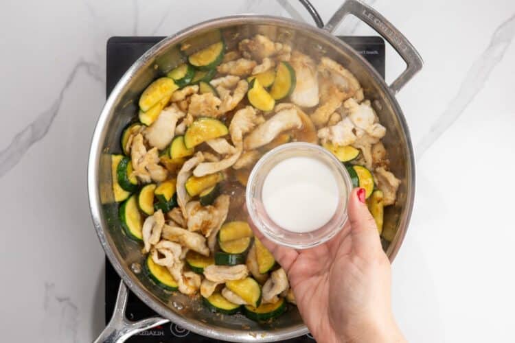 corn starch slurry in a small bowl, held over a pan of chicken stir fry with zucchini.