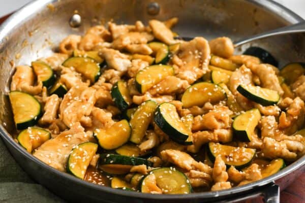 closeup of a skillet full of stir fry chicken and zucchini, garnished with sesame seeds.