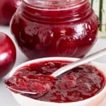 Spoonful of homemade plum jam over an open jar surrounded by fresh plums.