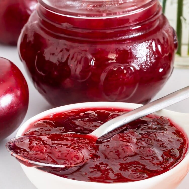 Spoonful of homemade plum jam over an open jar surrounded by fresh plums.