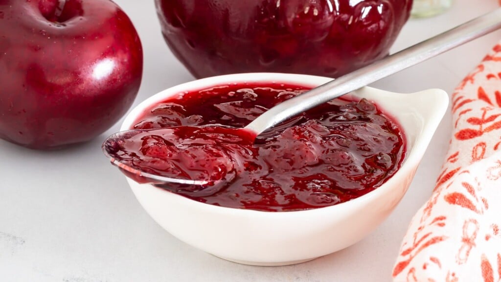 Chunky homemade plum jam in a white bowl with fresh red plums and a spoon.