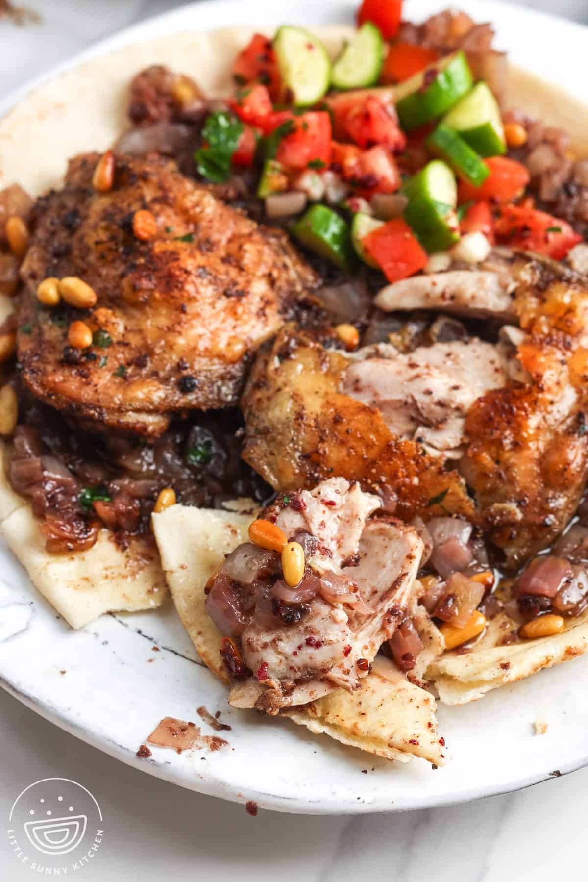 Cut-open sumac chicken thighs with onions, pine nuts, pita bread, and tomato cucumber salad.