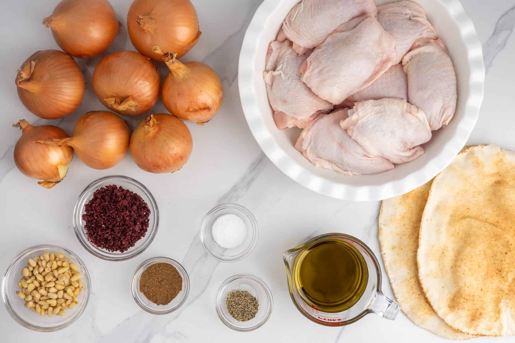 Ingredients for sumac chicken thighs, including bone-in chicken, onions, sumac, olive oil, pine nuts, spices, and pita bread.