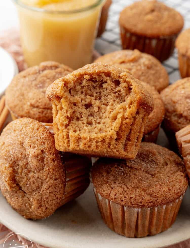 a pile of applesauce muffins topped with cinnamon sugar. One has a bite taken out to show the fluffy texture.