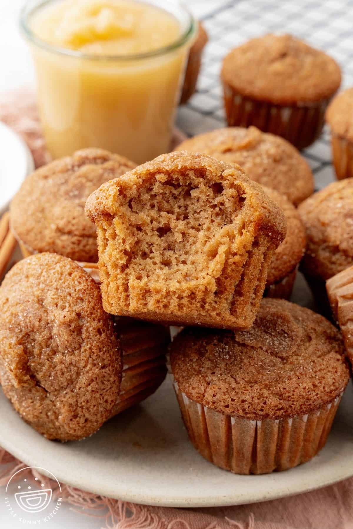 a pile of applesauce muffins topped with cinnamon sugar. One has a bite taken out to show the fluffy texture.