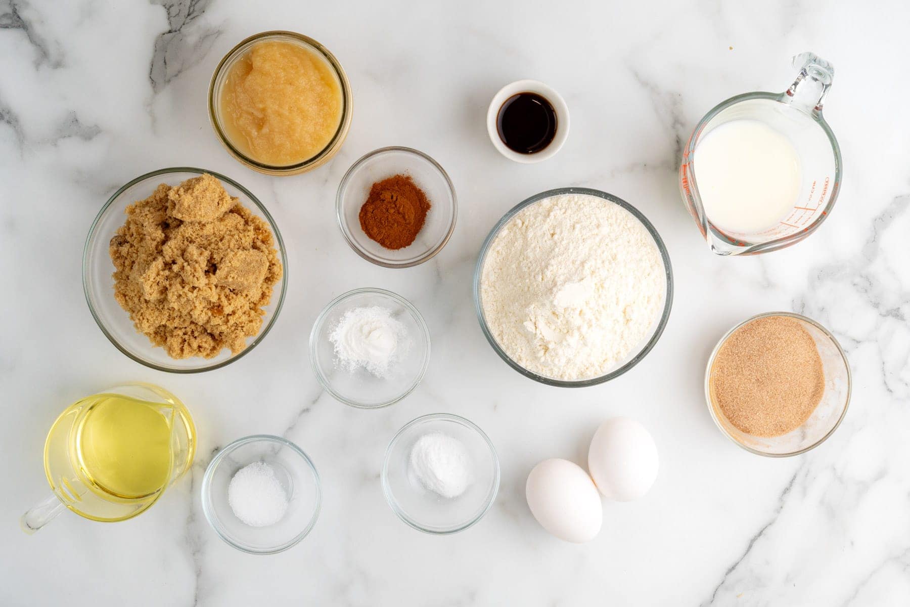 Ingredients for making muffins with applesauce, measured into small bowls, arranged on a marble counter