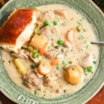 overhead view of a large bowl of rich, creamy chicken stew, set on a table. There is a fluffy dinner roll on the edge of the bowl.