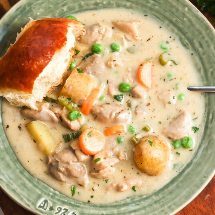 overhead view of a large bowl of rich, creamy chicken stew, set on a table. There is a fluffy dinner roll on the edge of the bowl.