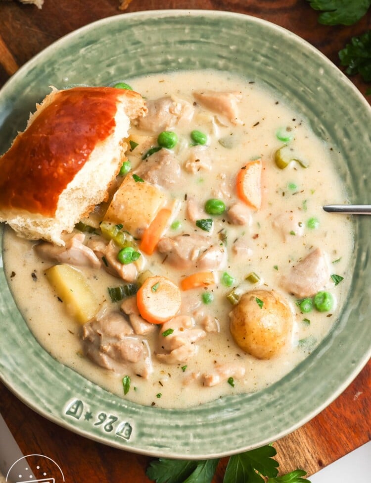 overhead view of a large bowl of rich, creamy chicken stew, set on a table. There is a fluffy dinner roll on the edge of the bowl.