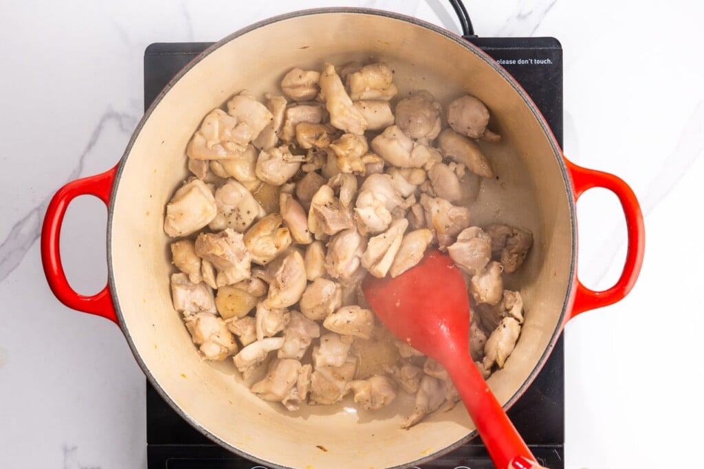 diced chicken thigh meat cooking in a dutch oven, stirred with a red spoon.
