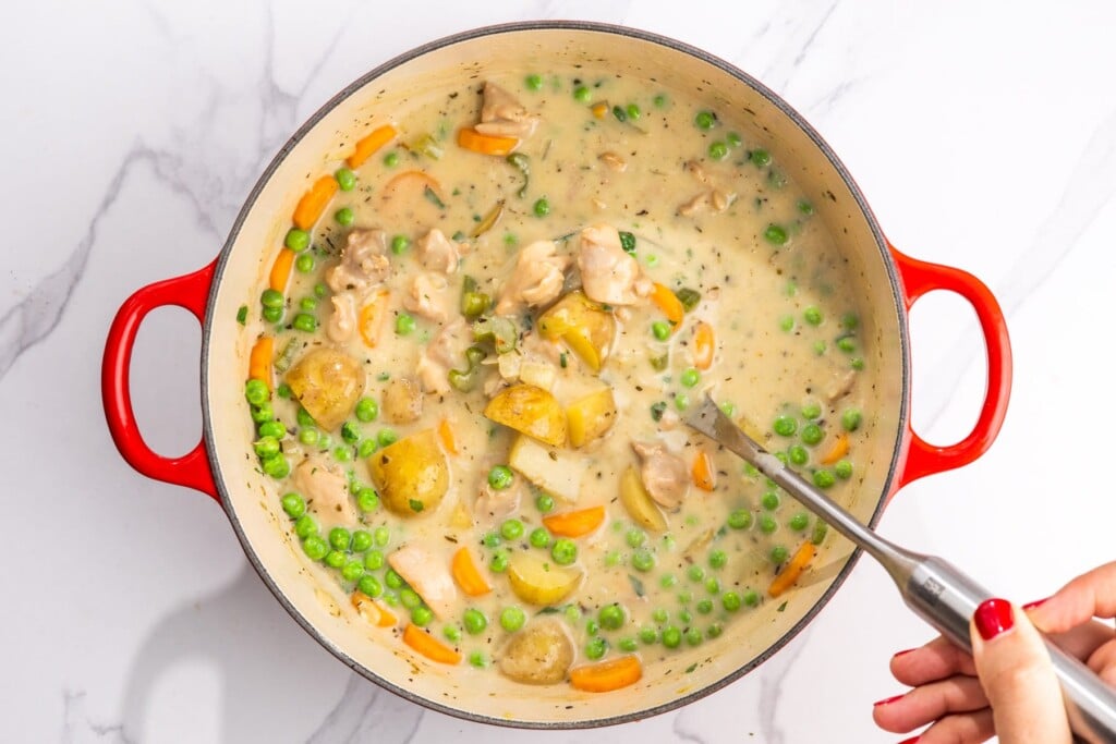 creamy, thickened chicken stew with potatoes, carrots, and peas, in a red dutch oven, stirred.